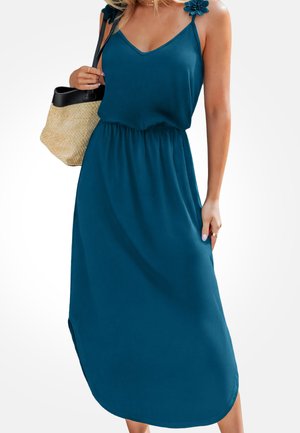 Frau trägt ein ärmelloses blaues Midikleid mit Blütendetails an den Trägern und hält eine beige-schwarze gewebte Handtasche auf einer Schulter.
