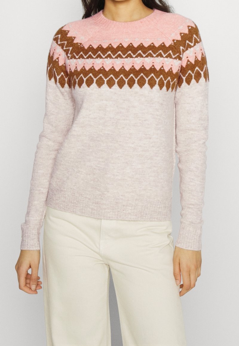 Femme portant un pull rose clair avec des motifs géométriques marron et roses sur la poitrine et les épaules, associé à un pantalon couleur crème.
