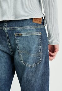 Niebieskie dżinsy z tylną kieszenią z detalami przeszycia. Nad pasem widoczna jest brązowa skórzana metka z napisem "Lee".