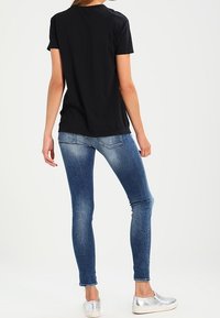 Mujer vestida con jeans ajustados azul desgastados, camiseta negra de manga corta y zapatos plateados tipo slip-on, de pie con la espalda hacia adelante.