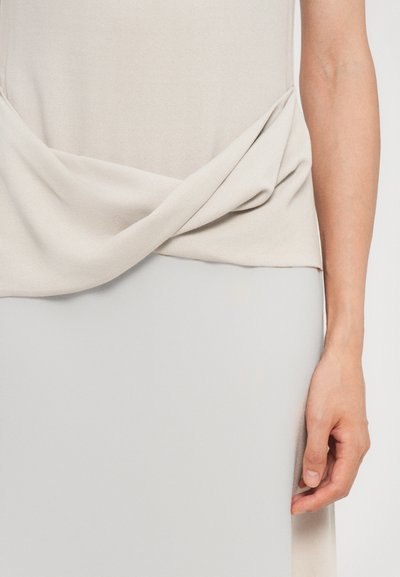 Beige top with a twisted front design, made of soft fabric. Smooth texture; paired with a light gray skirt, showcasing a clean, minimalist style.
