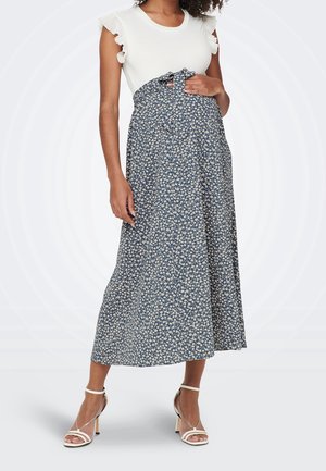 Kvinde iført hvid top uden ærmer med flæser, blå midilængde nederdel med højtaljede blomsterprint og hvide åbne højhælede sko med stropper.