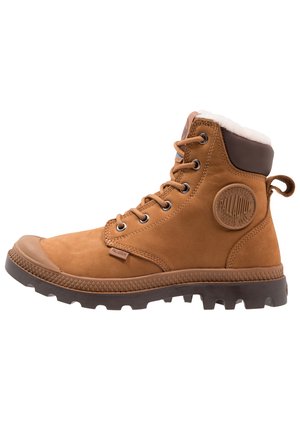 Brown suede ankle boot with a padded collar, rubber sole, metal eyelets, and a logo patch on the side. Textured grip and reinforced toe.