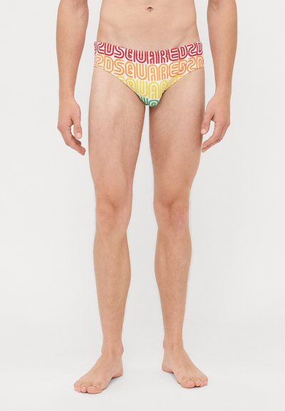 Modello maschile che indossa slip da bagno multicolori con marchio Dsquared2, in piedi a piedi nudi contro uno sfondo bianco semplice.