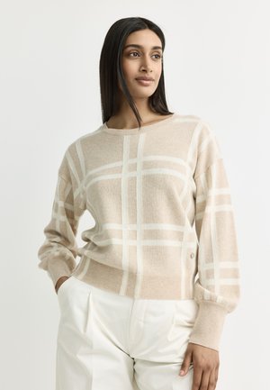 Woman wearing beige crewneck sweater with white plaid pattern and white high-waist pants, standing with one hand in pocket against plain background.