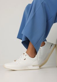 Baskets de sport blanches avec une finition texturée, des accents dorés sur les lacets et une semelle en caoutchouc beige, assorties à un pantalon large bleu.