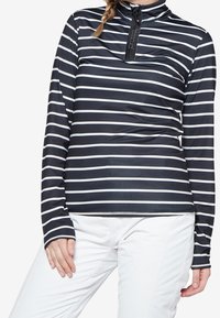 Donna che indossa un top a strisce navy e bianche a maniche lunghe con zip a quarto e pantaloni bianchi, in posa con una mano appoggiata sulla coscia.