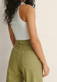 Weißes, kurz geschnittenes Tanktop und olivegrüne, hochtaillierte Hose mit sichtbaren Nähten und zwei Gesäßtaschen. Lockige Haare sind teilweise sichtbar.