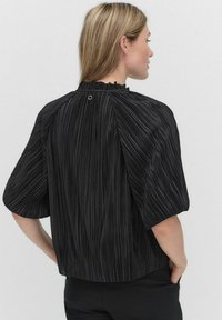 Sort kortærmet bluse med lodrette striber, der har en høj halsudskæring og en rynket detalje bagpå, sikret med en cirkulær pynt.