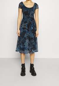 Robe noire avec des motifs floraux et paisley bleus, à manches courtes, avec un décolleté arrondi et une longueur au genou. Portée avec des bottines noires.