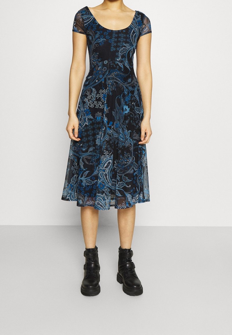 Robe noire avec des motifs floraux et paisley bleus, à manches courtes, avec un décolleté arrondi et une longueur au genou. Portée avec des bottines noires.