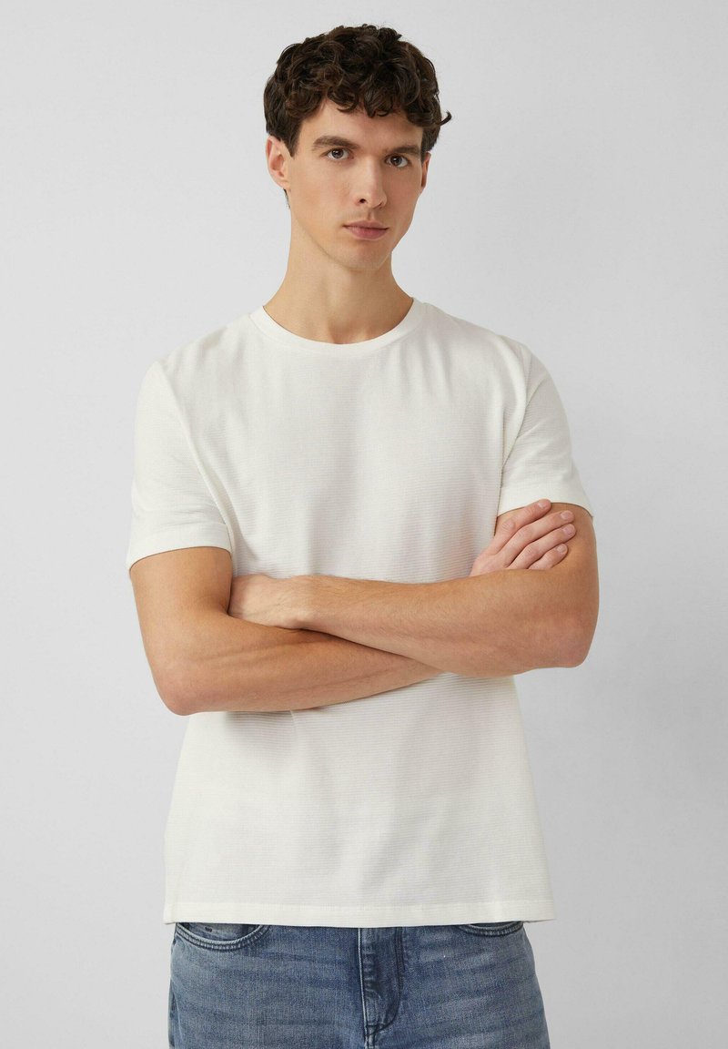 Camiseta blanca de manga corta con cuello redondo, hecha de tela texturizada. El ajuste es relajado, con un dobladillo recto. Se lleva con vaqueros azul claro.