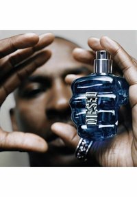 Sininen, nyrkin muotoinen parfyympullo, jossa on metallinen hopeanvärinen korkki ja paksut "DIESEL"-kirjaimet. Taustalla osittain näkyvät kädet.