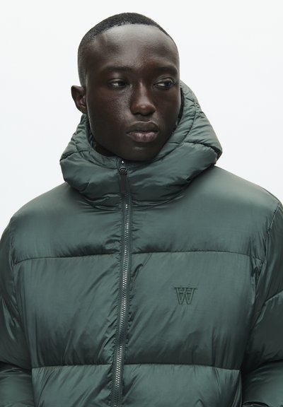 Homme portant une doudoune vert foncé avec un col haut, regardant sur le côté devant un fond clair.