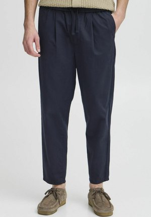 Mand iført marineblå bukser med snøre, beige strikket top og brune ruskindssko med snørebånd, stående med den ene hånd i lommen.