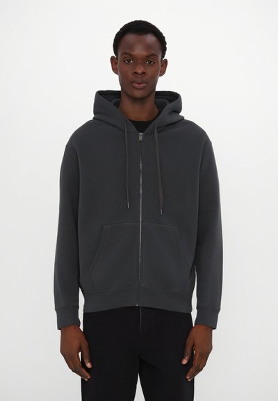 Dark gray zip-up hoodie with a drawstring hood, front pockets, ribbed cuffs, and a smooth texture. The model wears it with black pants.