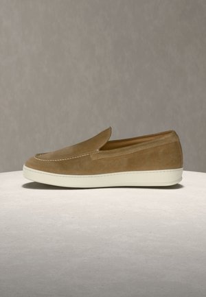 Zapato tipo mocasín de gamuza marrón con suela de goma blanca, mostrado de perfil sobre una superficie clara y texturizada.