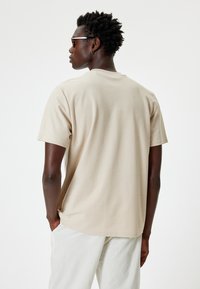 Kort beige T-shirt, blødt stof, rund halsudskæring, korte ærmer, afslappet pasform, båret med lyse bukser, ensartet design, ingen mønstre.