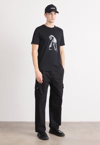 Black t-shirt featuring a graphic of a dog, paired with black cargo pants and black shoes. A black cap with "HUGO" is worn. Neutral background.