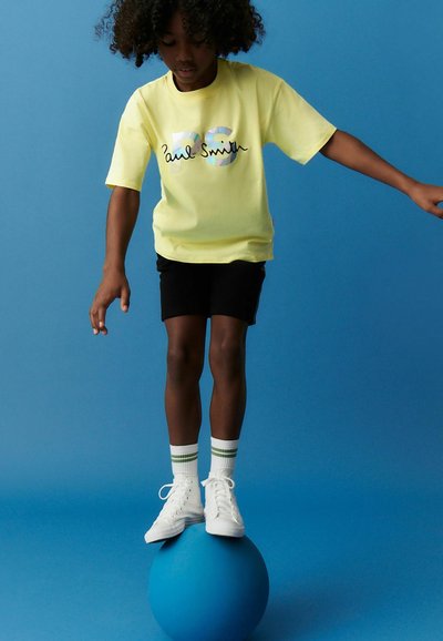 Camiseta amarilla de manga corta con el logo "Paul Smith", pantalones cortos negros, zapatillas blancas y calcetas a rayas verdes, equilibrándose sobre una pelota azul.
