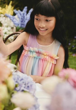 Noor tüdruk naeratab, istudes õues laua taga, kandes pastelseid vikerkaarevärve kleiti, ümbritsetud värvikatest lilleõitest.