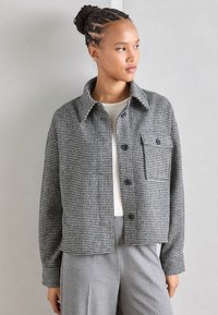 Black and white checked jacket with a collar, front buttons, and a chest pocket. Worn over a white top, paired with grey trousers.