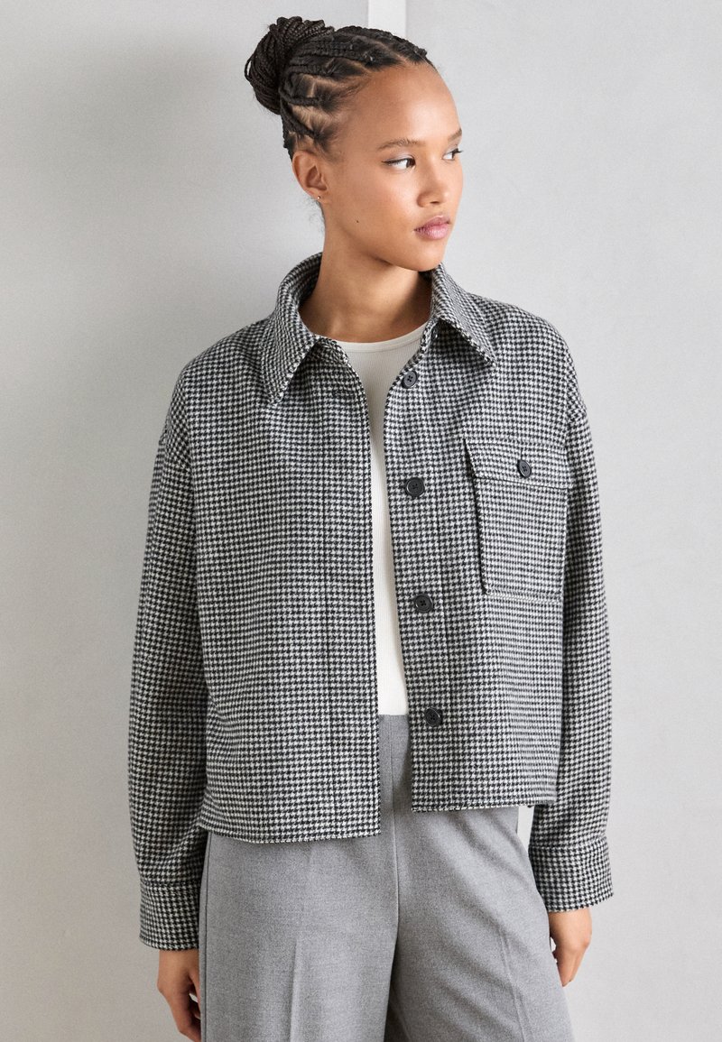 Black and white checked jacket with a collar, front buttons, and a chest pocket. Worn over a white top, paired with grey trousers.