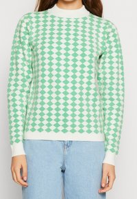 Maglione a motivi a diamante verdi e bianchi con scollo rotondo e polsini a coste, realizzato in morbido materiale lavorato a maglia, abbinato a jeans azzurri.