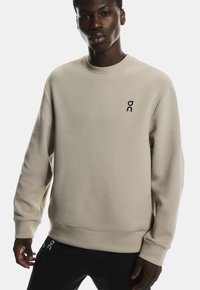 Hanorac bej cu guler rotund, manșete și tiv cu dungi, având un mic logo negru pe pieptul stâng. Textură a materialului moale, croială lejeră.