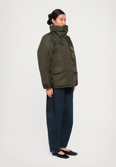Woman standing sideways wearing a dark green padded jacket, wide dark blue jeans, and black flat shoes on a plain white background.
