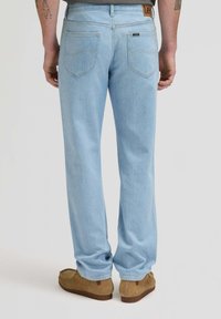 Hombre con vaqueros azul claro y zapatos de ante marrón, visto desde atrás sobre un fondo liso.