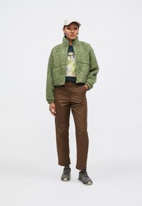 Zelená plyšová bunda na zip, grafická mikina, hnědé kalhoty a šedé turistické boty. Čepice má světlou neutrální barvu. Jednoduchý design.