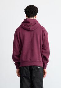 Felpa bordeaux con un cappuccio ampio, polsini e orlo a coste. Tessuto morbido al tatto. Toppa nera con logo su pantaloni scuri.