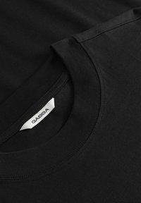 Gros plan sur un T-shirt noir à col rond montrant la couture côtelée du col et une étiquette de marque GABBA blanche à l’intérieur de l’encolure.