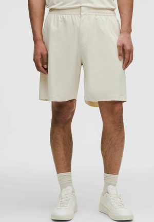 Mand iført cremefarvede shorts med elastik i taljen, matchende sokker og hvide sneakers, stående mod en ensfarvet baggrund.