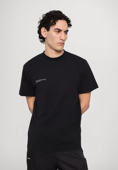 Tricou negru din bumbac, cu guler rotund, mâneci scurte și accent de text alb pe partea din față, purtat cu pantaloni negri.