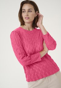 Pull en rose texturé avec un motif en diamants, encolure ronde et manches longues. Le tissu a une légère brillance, ajoutant de la profondeur à la couleur.
