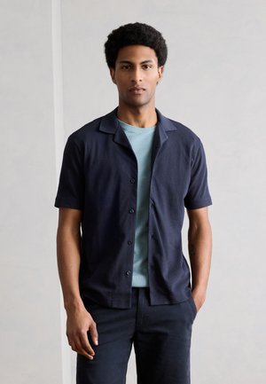 Νέος άνδρας με κοντά σγουρά μαλλιά, φορώντας έναν γαλάζιο T-shirt κάτω από ένα σκούρο μπλε πουκάμισο με κοντά μανίκια, στέκεται με το ένα χέρι στην τσέπη.