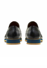 Une paire de chaussures de ville en cuir noir avec des détails de brogue, semelles superposées bleu et taupe, montrées de l'arrière sur un fond blanc.