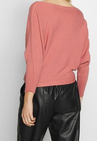 Maglione rosa con coste, scollo ampio e maniche lunghe, abbinato a pantaloni in pelle neri lucidi a vita alta.