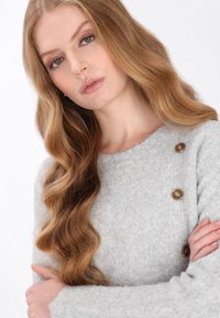 Lys grå sweater lavet af blødt materiale, med rund halsudskæring, tekstureret mønster og to dekorative brune knapper på skulderen.