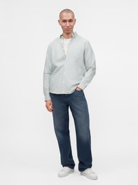 Jeune homme debout, une main dans la poche, portant une chemise à rayures claires boutonnée, un t-shirt blanc, un jean bleu et des baskets blanches sur fond uni.