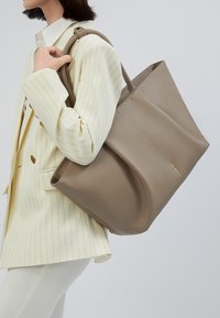 Vrouw in een crèmekleurige pinstripe blazer en witte broek die een grote taupe leren tote tas over haar schouder houdt.