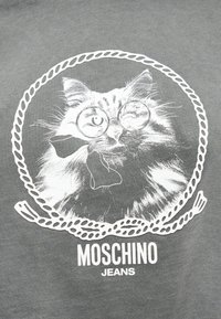Αφράτη γάτα που φοράει στρογγυλά γυαλιά και παπιγιόν, πλαισιωμένη από έναν κύκλο σχοινιού, με το κείμενο «Moschino Jeans» από κάτω σε γκρι ύφασμα.