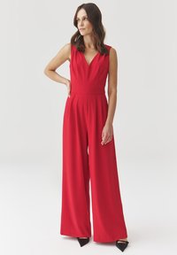 Rode mouwloze jumpsuit met een V-hals, brede pijpen en een getailleerde taille, gemaakt van een gladde stof. Gekozen met zwarte puntige hakken.