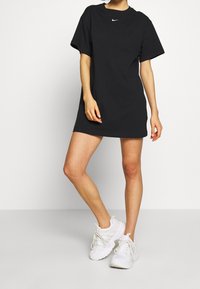 Svart bomullst-shirtklänning med korta ärmar; har en liten vit Nike-logotyp på bröstet. Matchad med vita sneakers.
