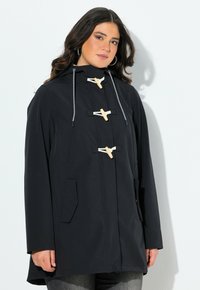 Kvinna som bär en svart huva med träknappar och vita repöglor, tillsammans med mörkgrå jeans mot en vit bakgrund.