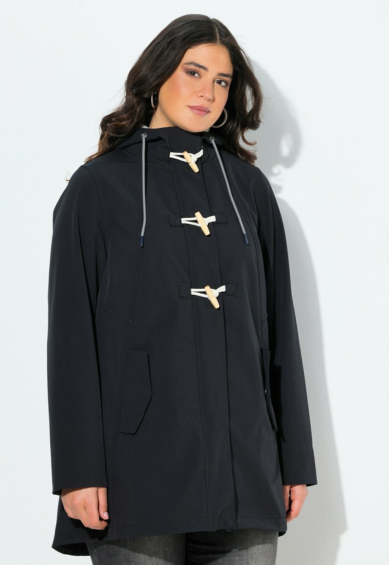 Kvinna som bär en svart huva med träknappar och vita repöglor, tillsammans med mörkgrå jeans mot en vit bakgrund.