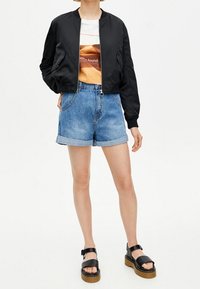 Černá saténová bomber bunda, bílý tričko s oranžovým designem, denimové šortky a černé sandály s platformou, představující ležérní outfit.