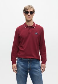 Bordeaux langarm poloshirt met witte kraagafwerking en merkpatch, gecombineerd met blauwe spijkerbroek. Model draagt donkere zonnebril.